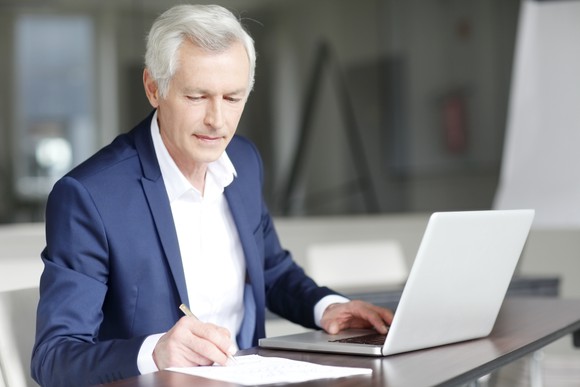 Gray-haired man in professional attire taking notes while typing on a laptop.