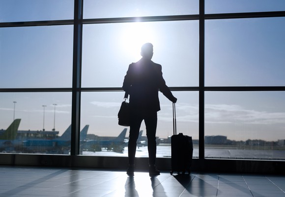 Man wearing shoulder bag and holding luggage looking out window of airport
