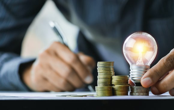 Illuminated lightbulb, resting on a stack of coins, with a hand writing on paper in the background.