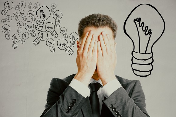 A man in a suit covering his eyes. On a wall behind him are drawings of light bulbs.