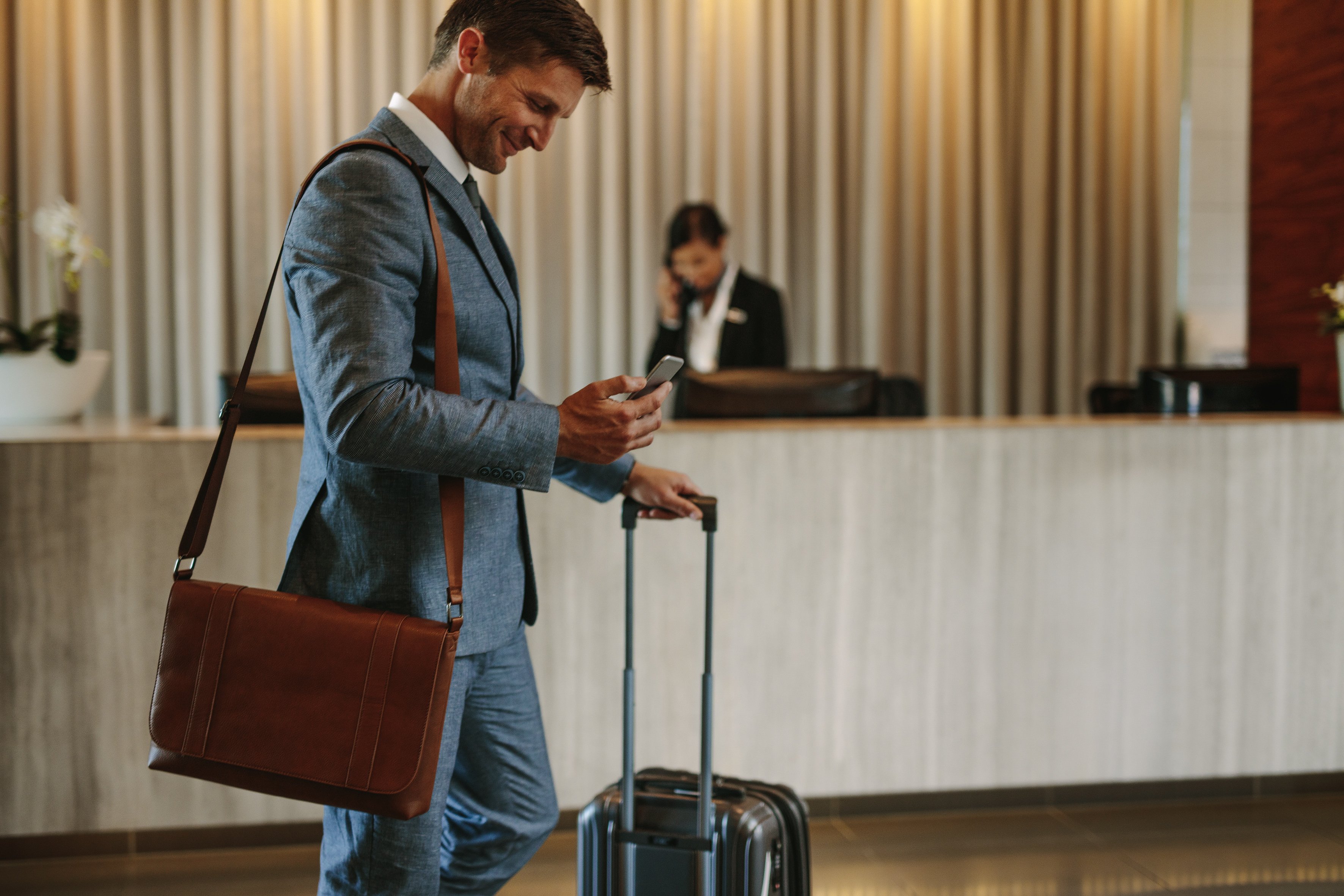 Man in suit checking phone in hotel lobby while holding wheeled suitcase and wearing shoulder bag