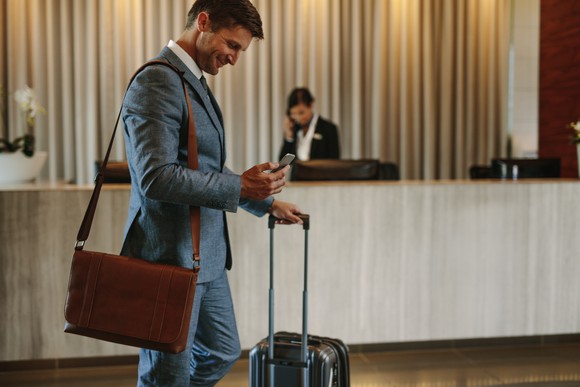 Man in suit checking phone in hotel lobby while holding wheeled suitcase and wearing shoulder bag