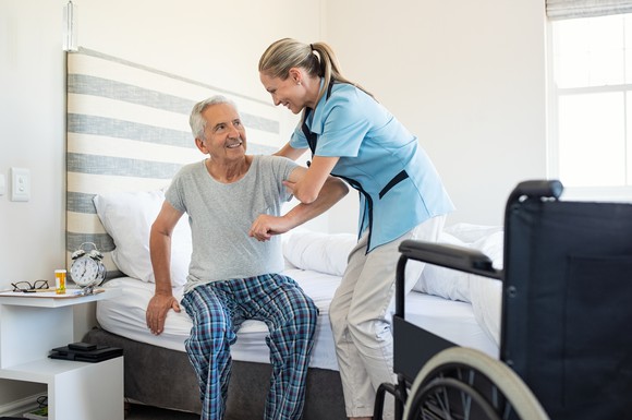 Woman in blue shirt and white pants helping senior man in pajamas out of bed