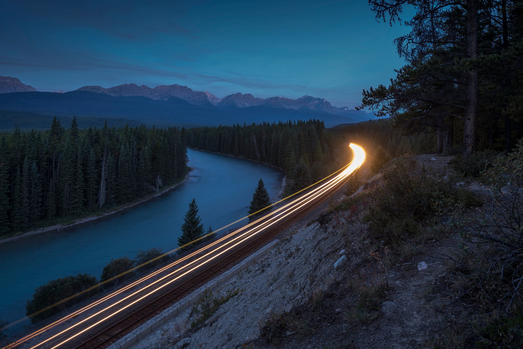 A locomotive's headlamps illuminate a river as rounds a bend by at night.