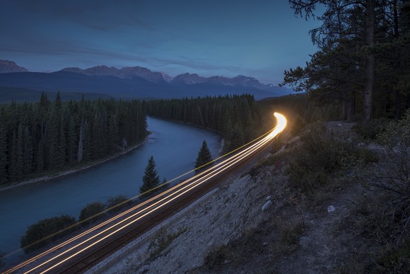 A locomotive's headlamps illuminate a river as rounds a bend by at night.