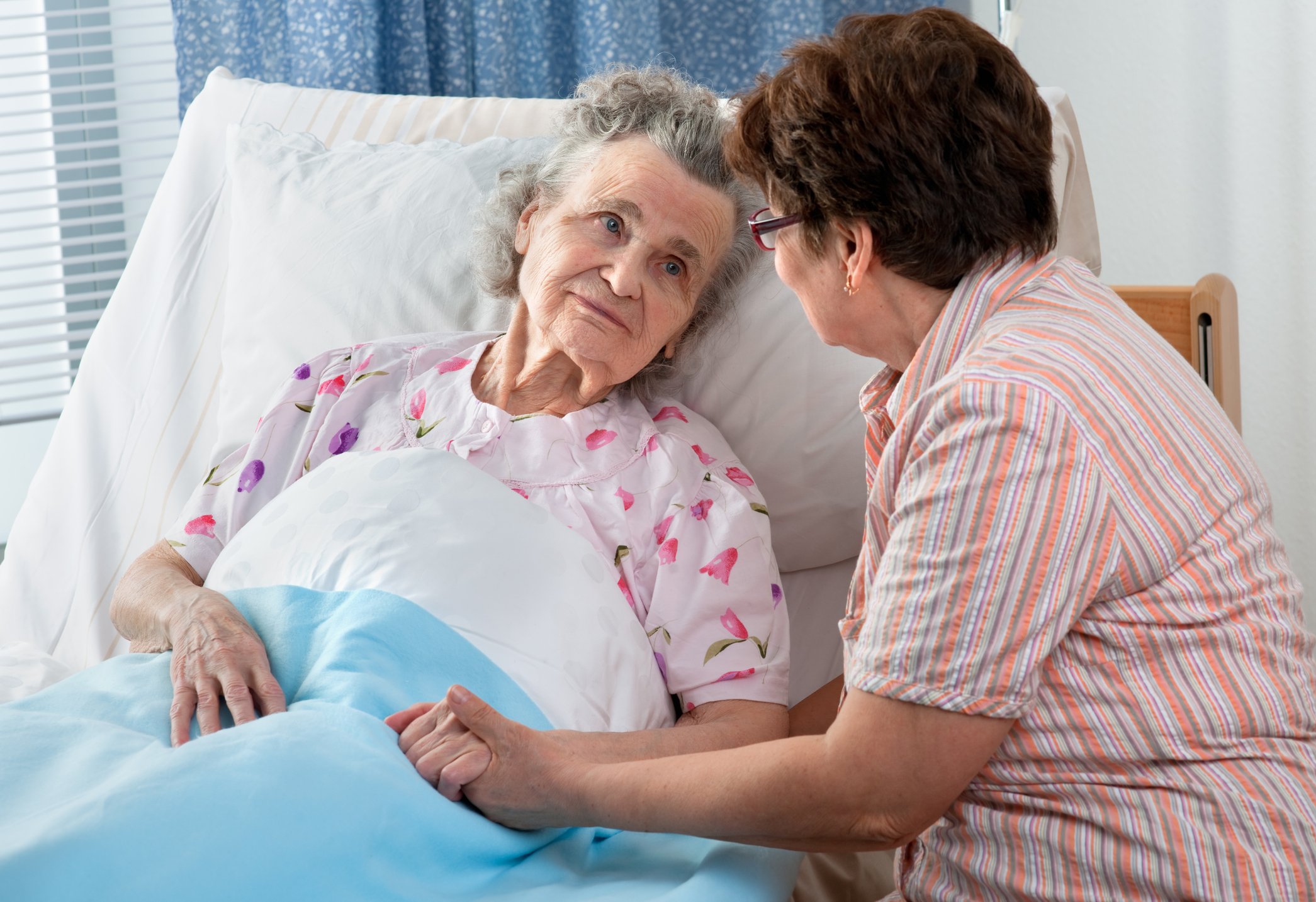 Older woman in bed while another woman sits with her, holding her hand