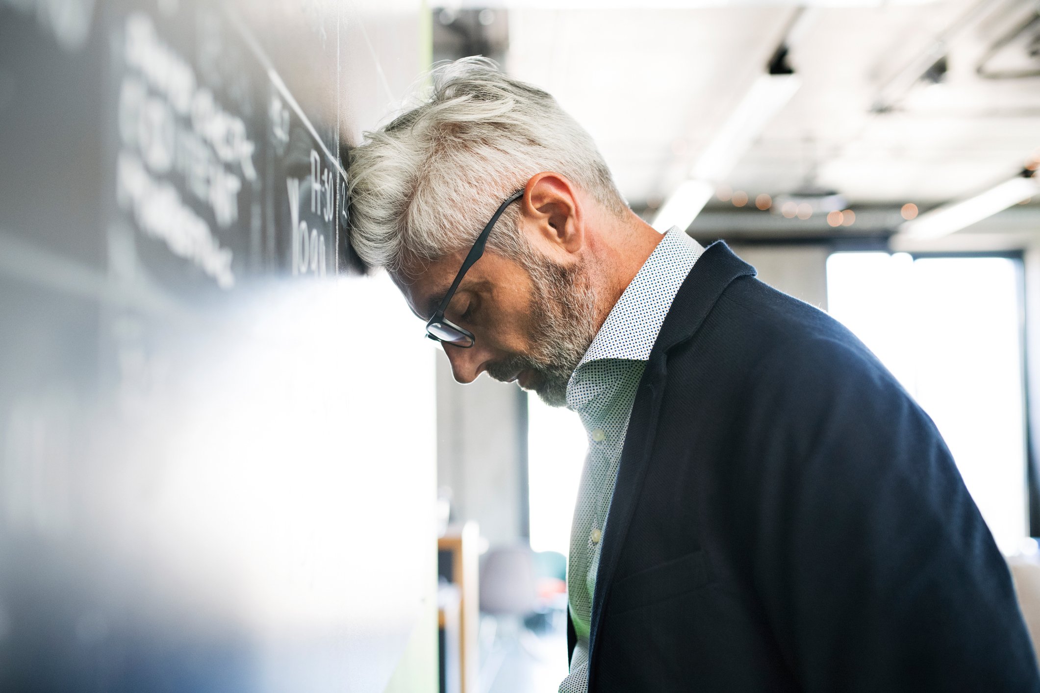 Gray-haired man in glasses, collared shirt, and suit jacket leaning head against wall