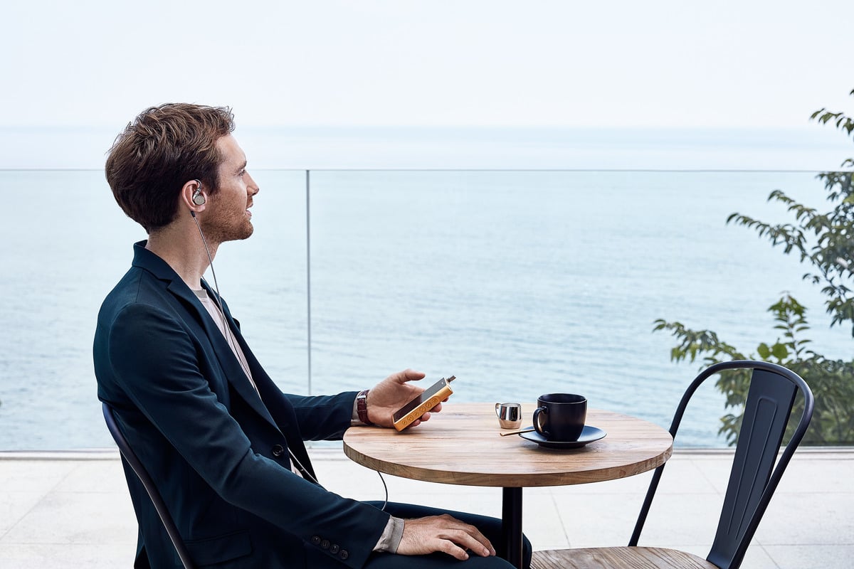 A man sitting at a small circular table with earbuds in holding a phone. The table is overlooking a large body of water.