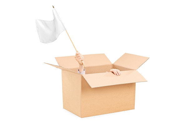 A man hiding in a cardboard box waves a white flag.