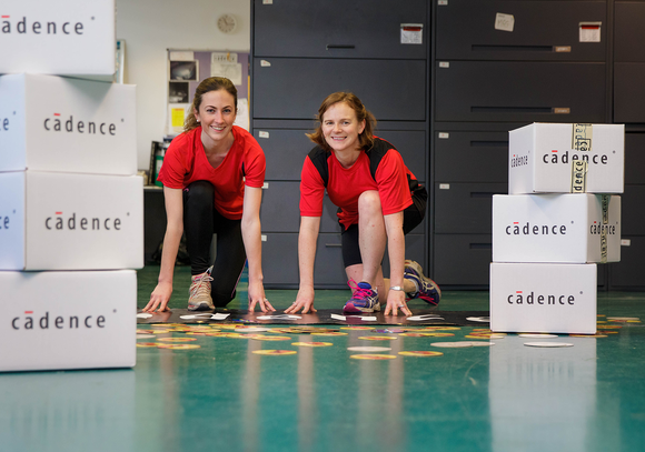 Two women in running outfits, in between two stacks of boxes with the Cadence logo on them