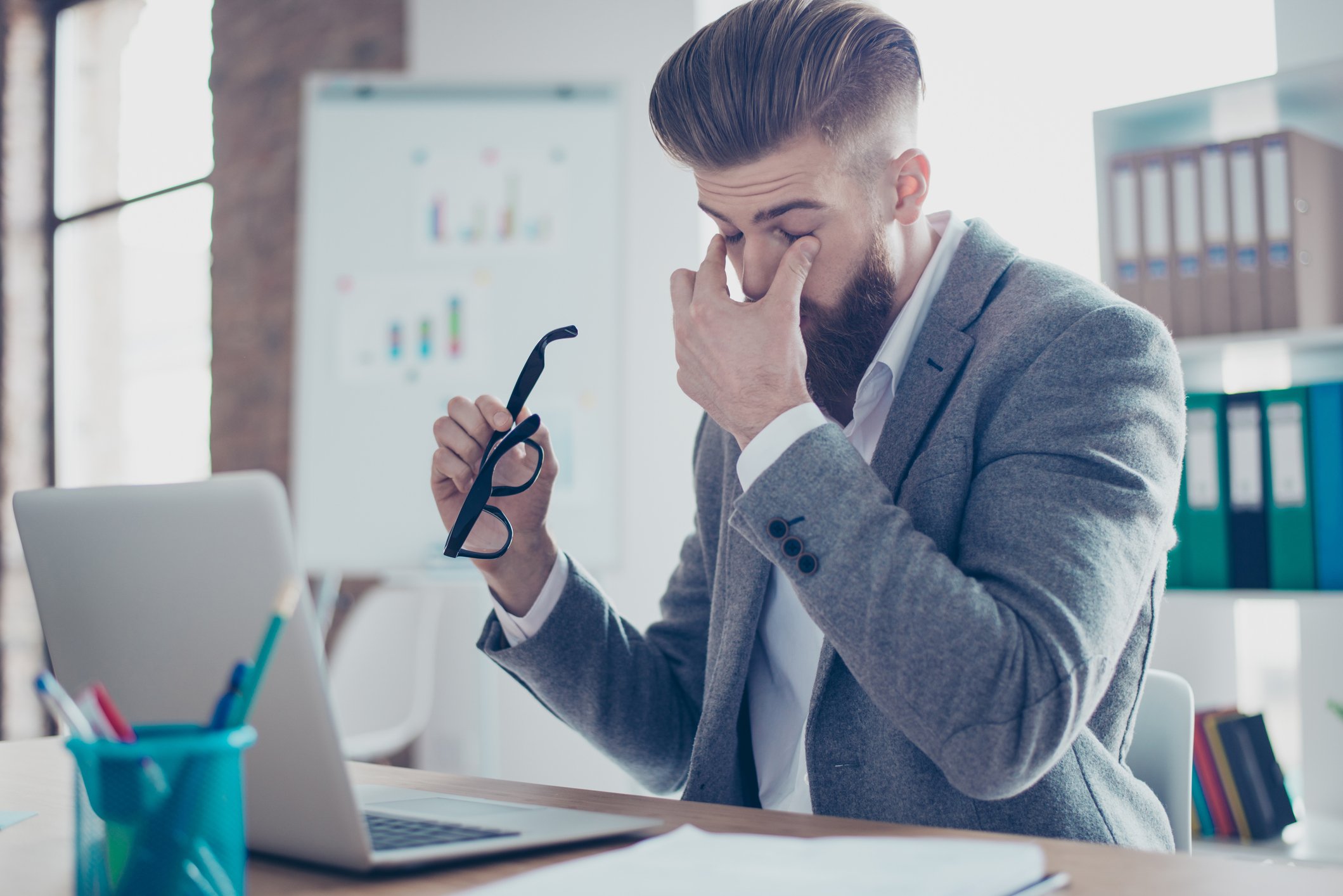 Man at laptop holding glasses with one hand and gripping around his nose with the other, as if tired or stressed