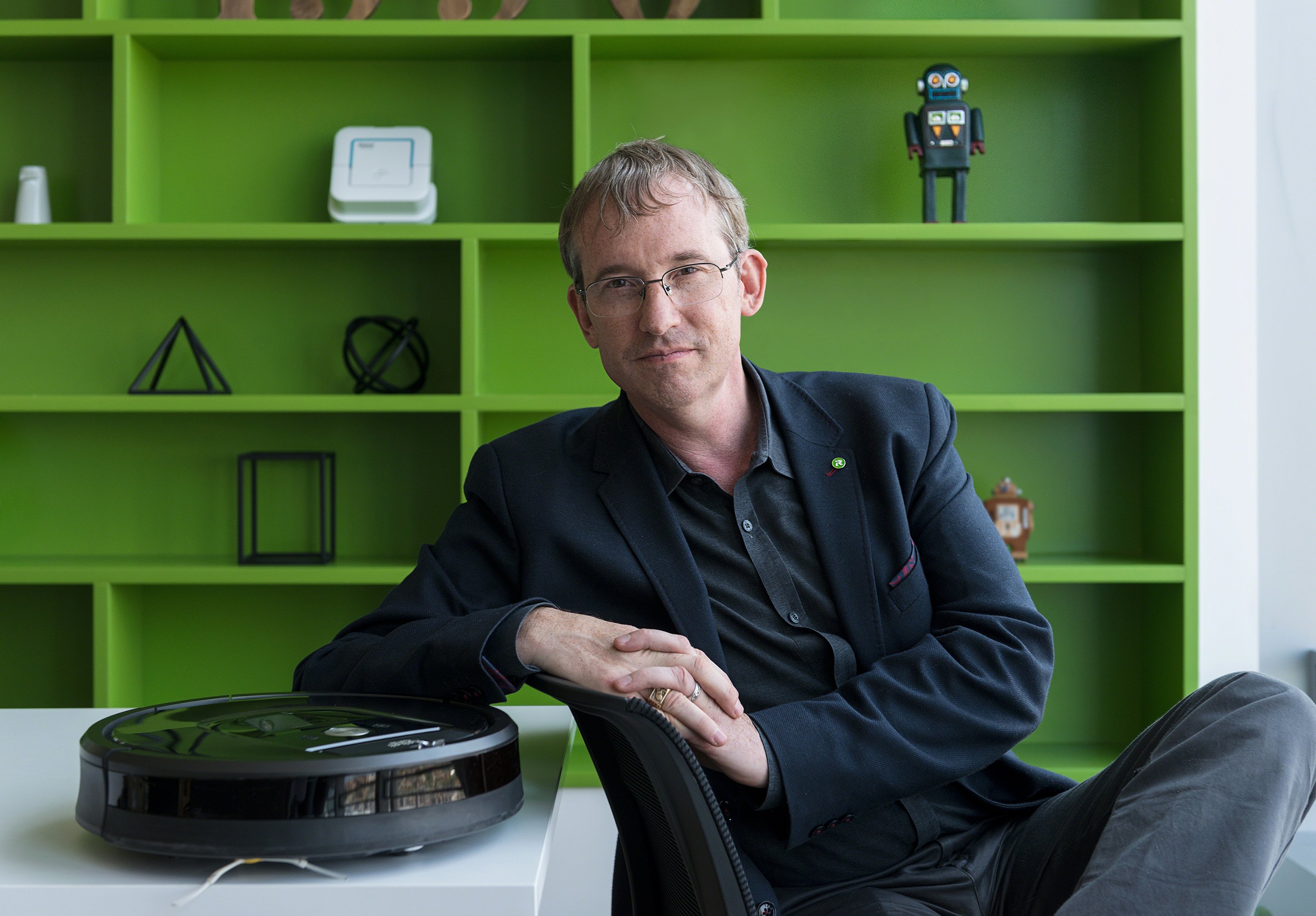 iRobot chairman, CEO, and co-founder Colin Angle sitting next to a Roomba with a green shelf in the background