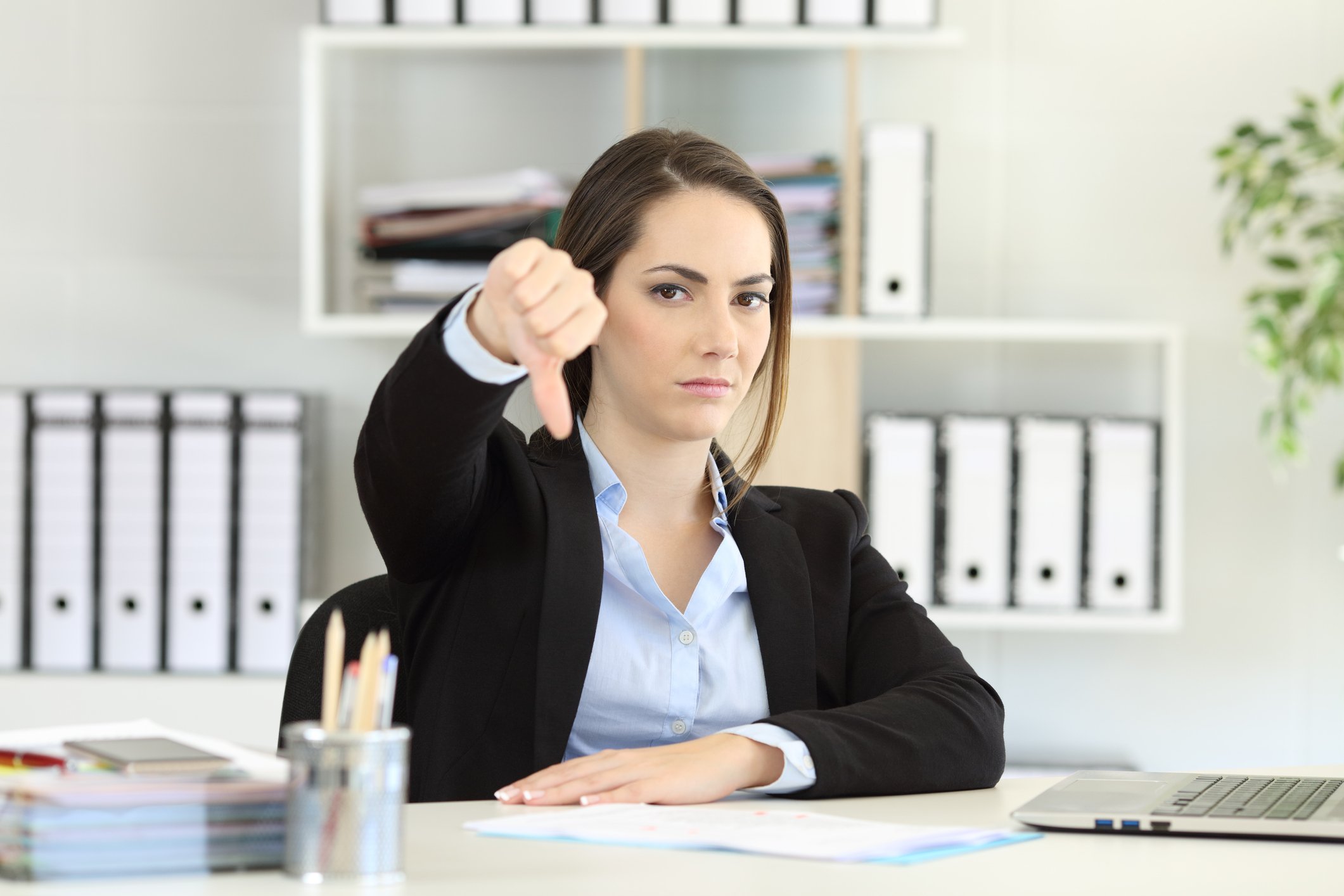 Woman in professional attire at desk making thumbs down sign