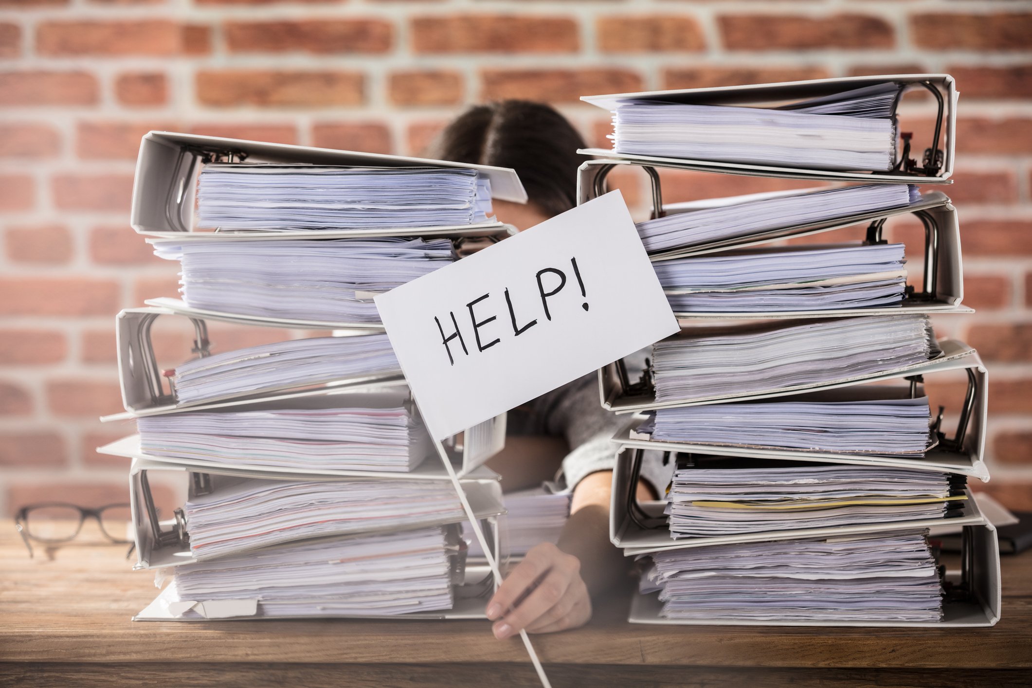 Woman sitting behind two large piles of binders holding sign reading help in between the piles