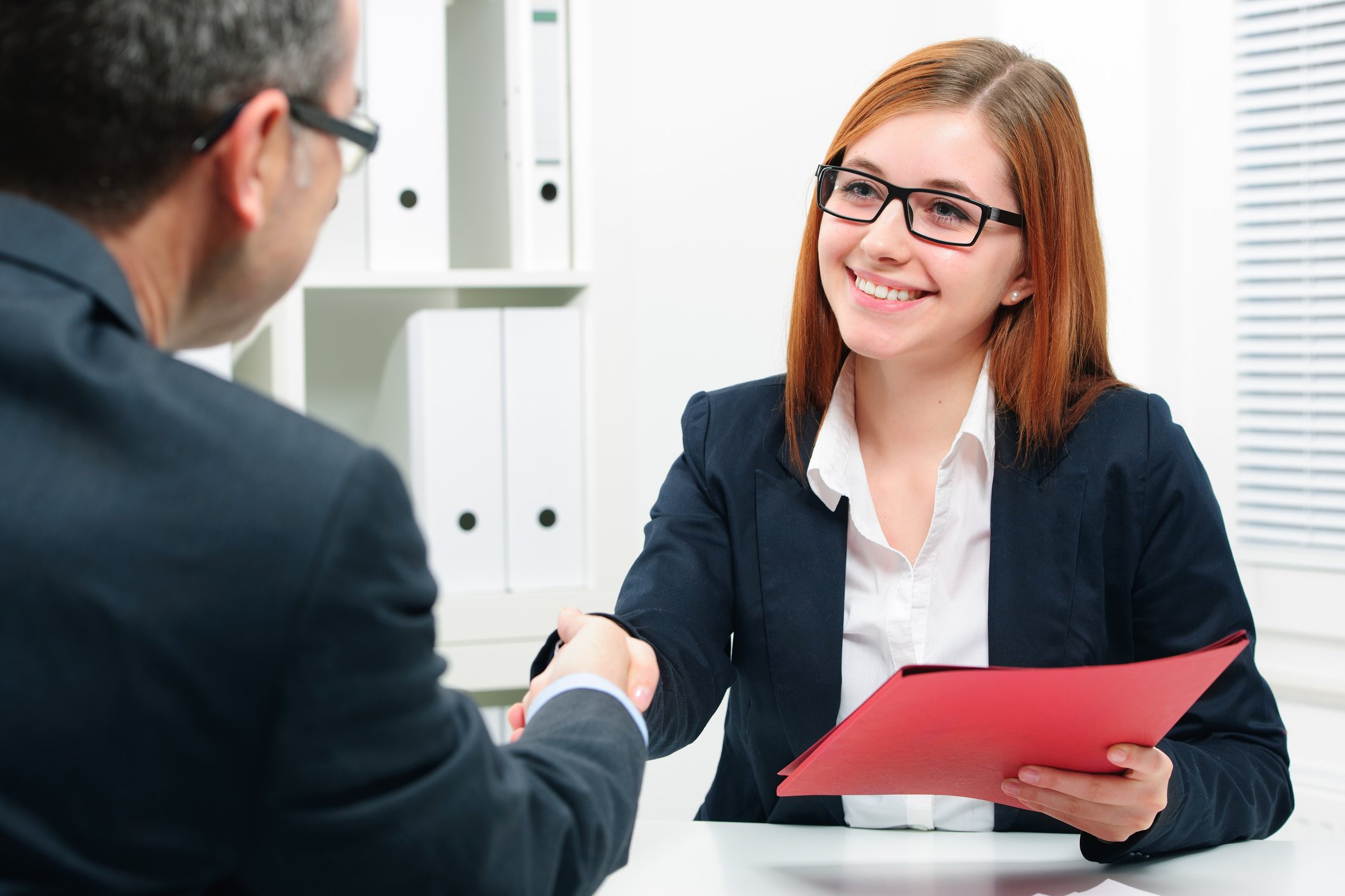 Professionally dressed woman holding red folder shaking hands with professionally dressed man seated across from her