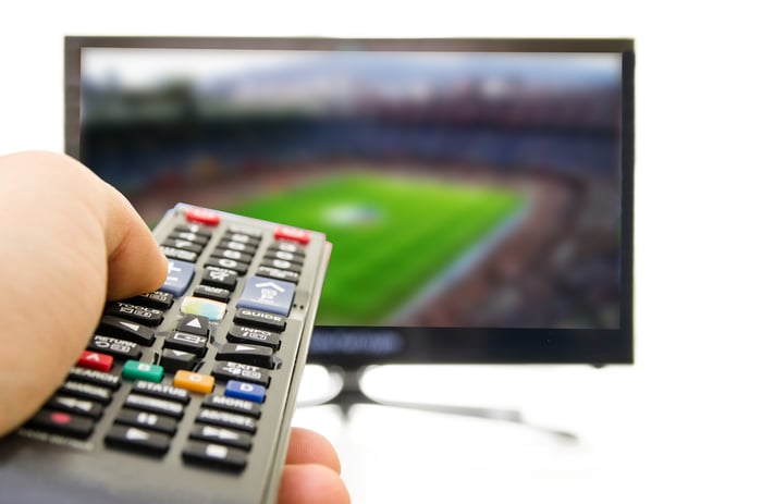 A TV in the background displaying a sporting event. A hand holding a remote control is displayed in the foreground.