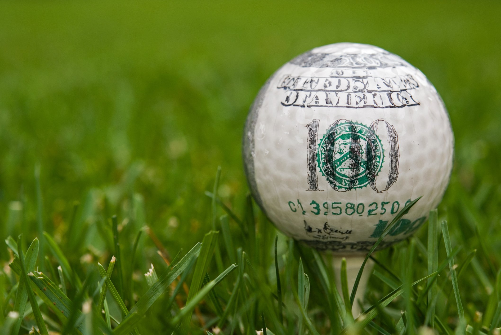 A golf ball with the markings of a hundred dollar bill on a tee in the grass.