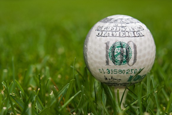 A golf ball with the markings of a hundred dollar bill on a tee in the grass.