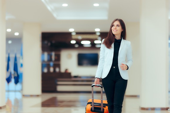 A businesswoman walks through a sparkling hotel lobby pulling a wheeled suitcase.