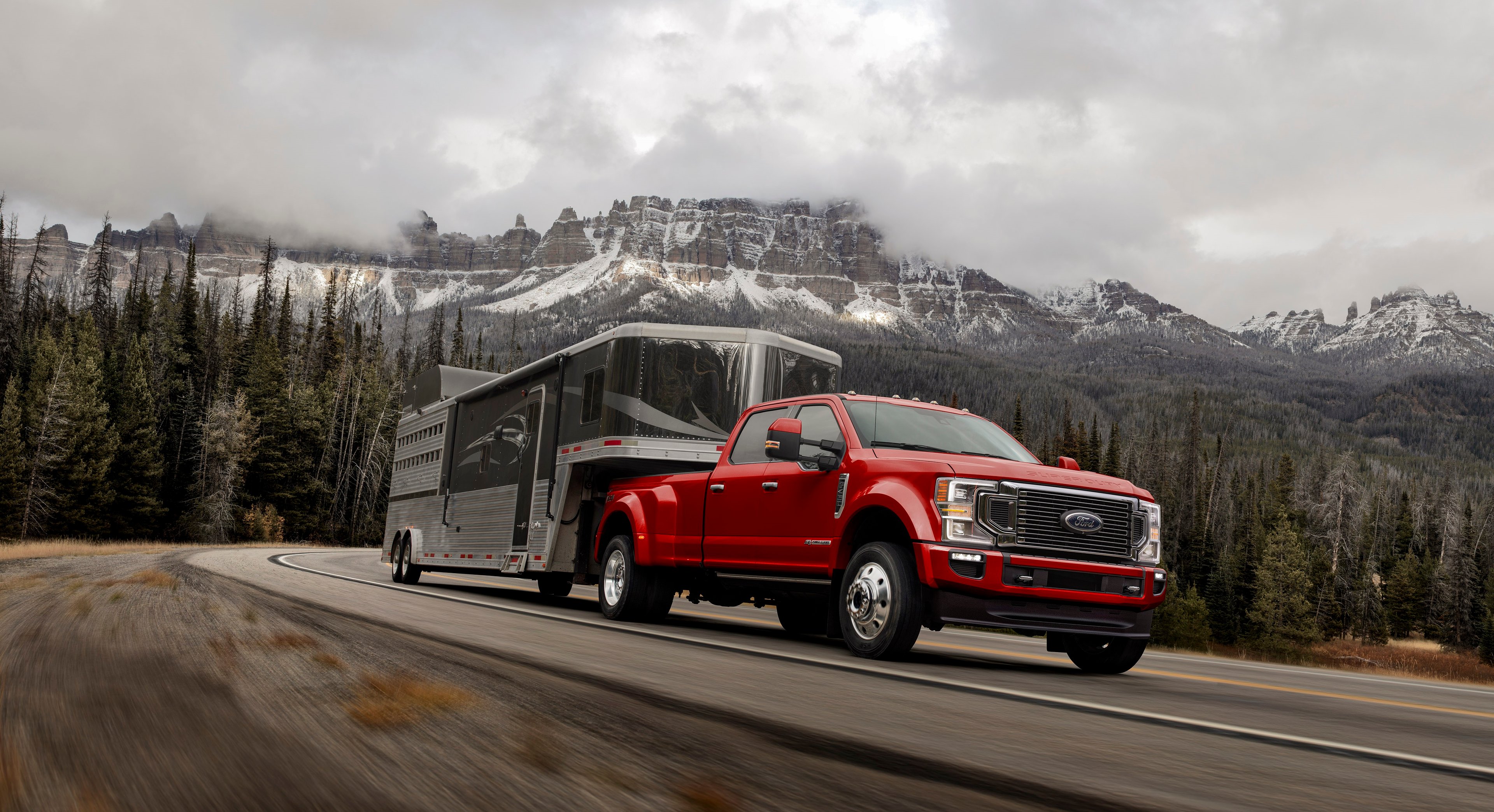 A red Ford F-450 hauling a trailer against a backdrop of snow-capped mountains and trees.