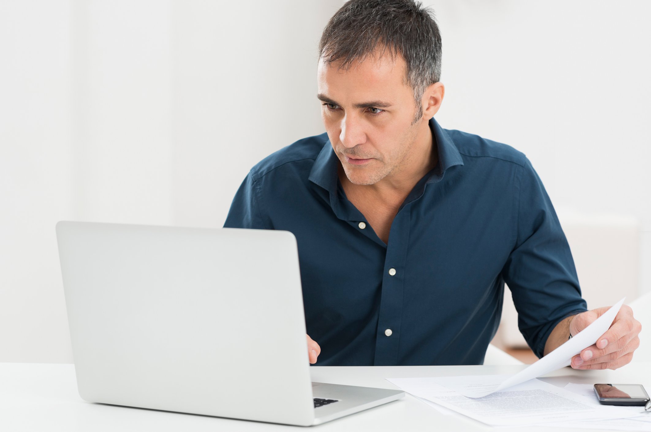 A man sits looking at a laptop monitor, while holding a document in one hand.
