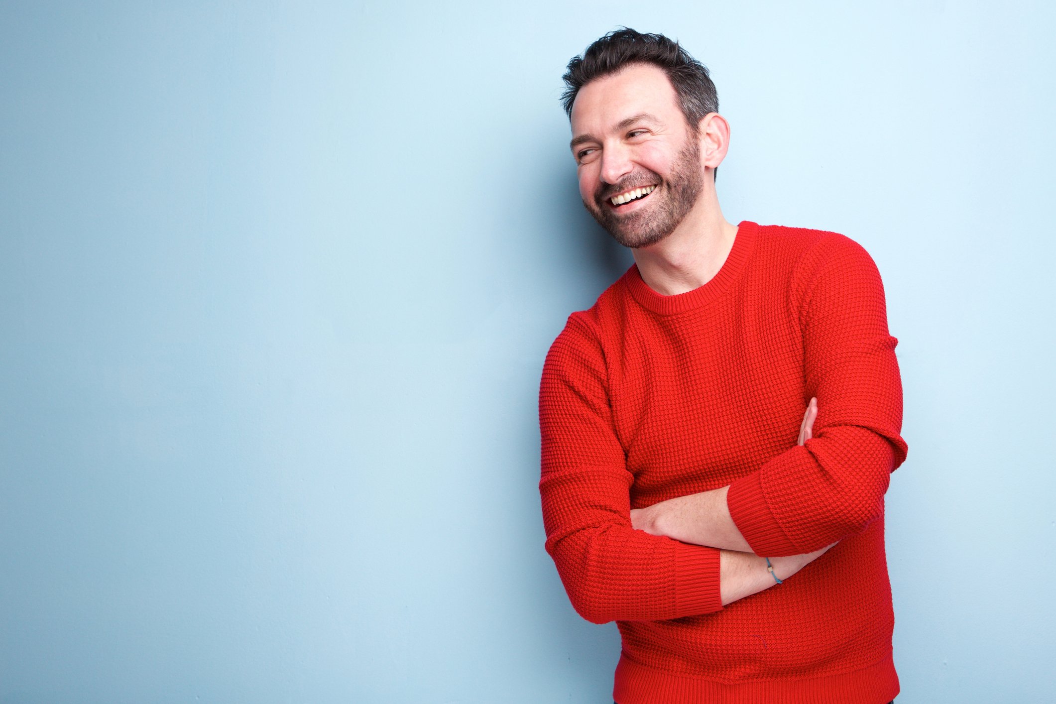 Smiling younger man in red shirt with arms crossed against blue background