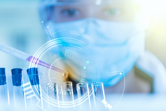 Person in lab mask looking at a dropper and test tubes.