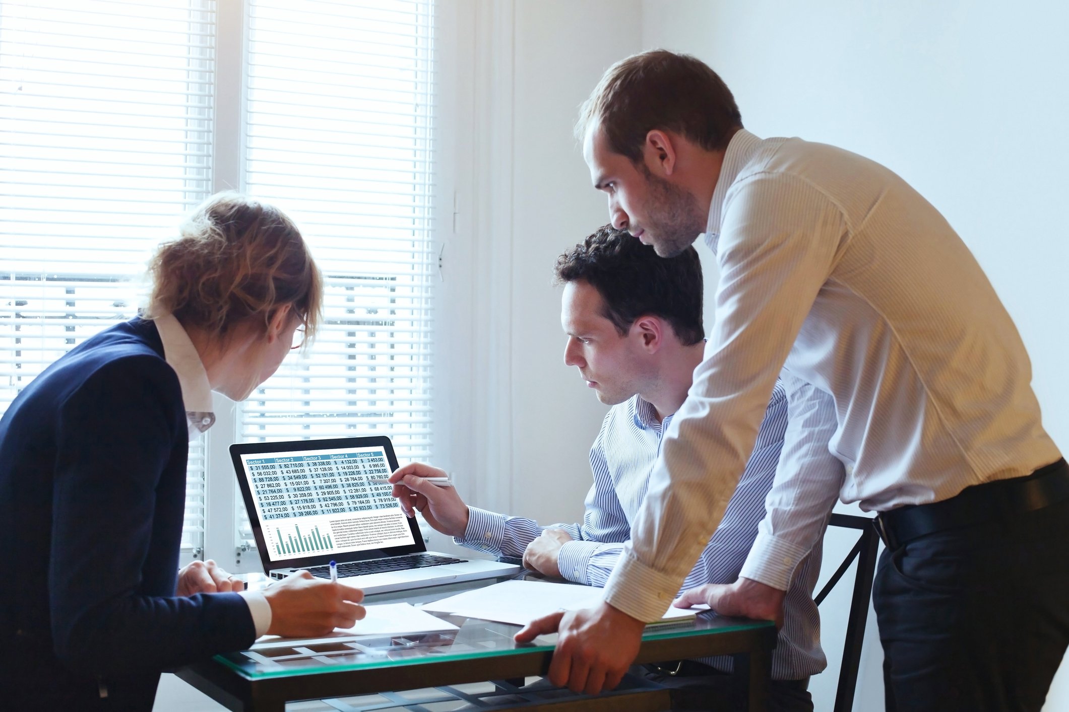 A group of three office workers gather around a computer displaying charts.