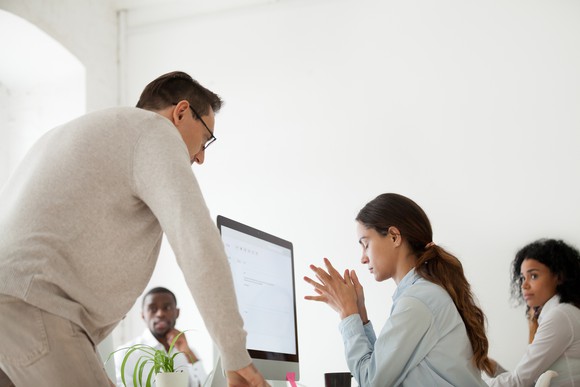 Man stands over a woman at a computer who has her eyes closed, as another man and woman look on.