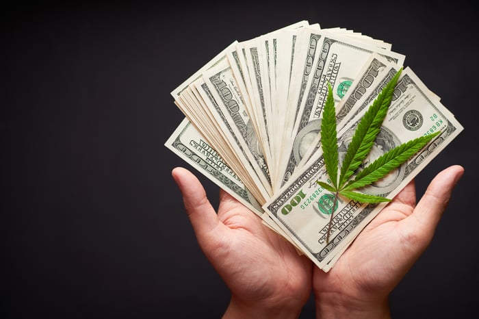 Hands holding $100 bills fanned out with a marijuana leaf on top.