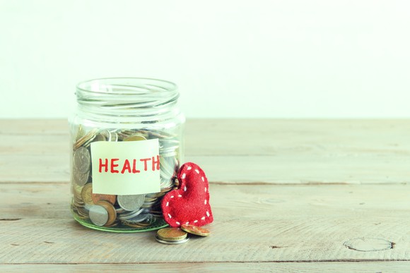 Glass jar filled with coins sitting on a wooden surface labeled health, with a felt red heart resting against it.