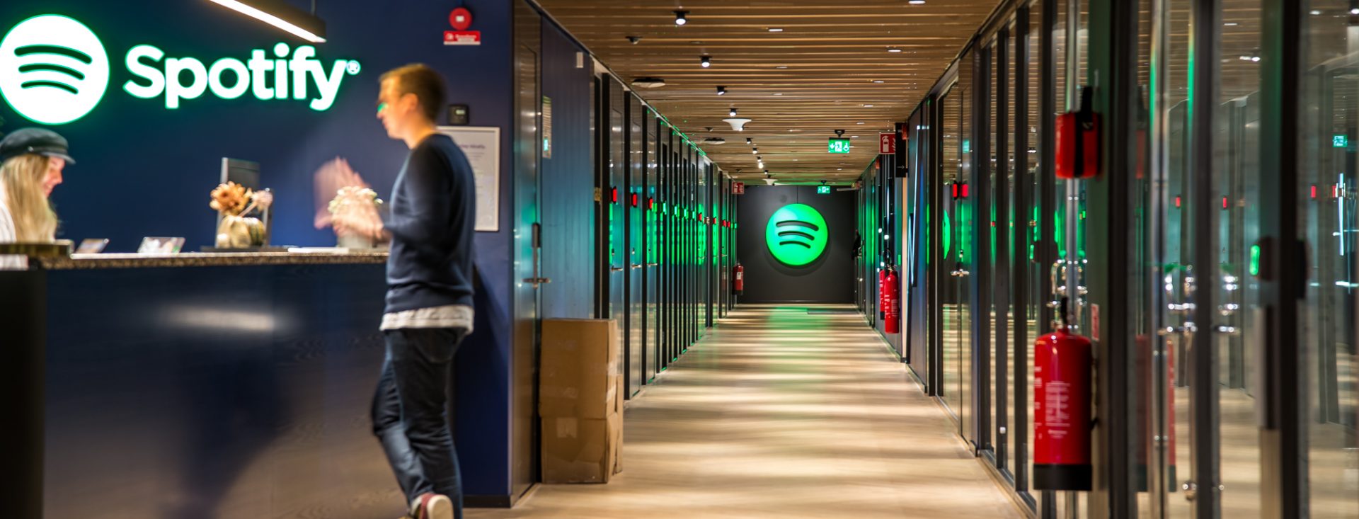 A man stands at a reception desk; the Spotify logo is visible down a long hallway.