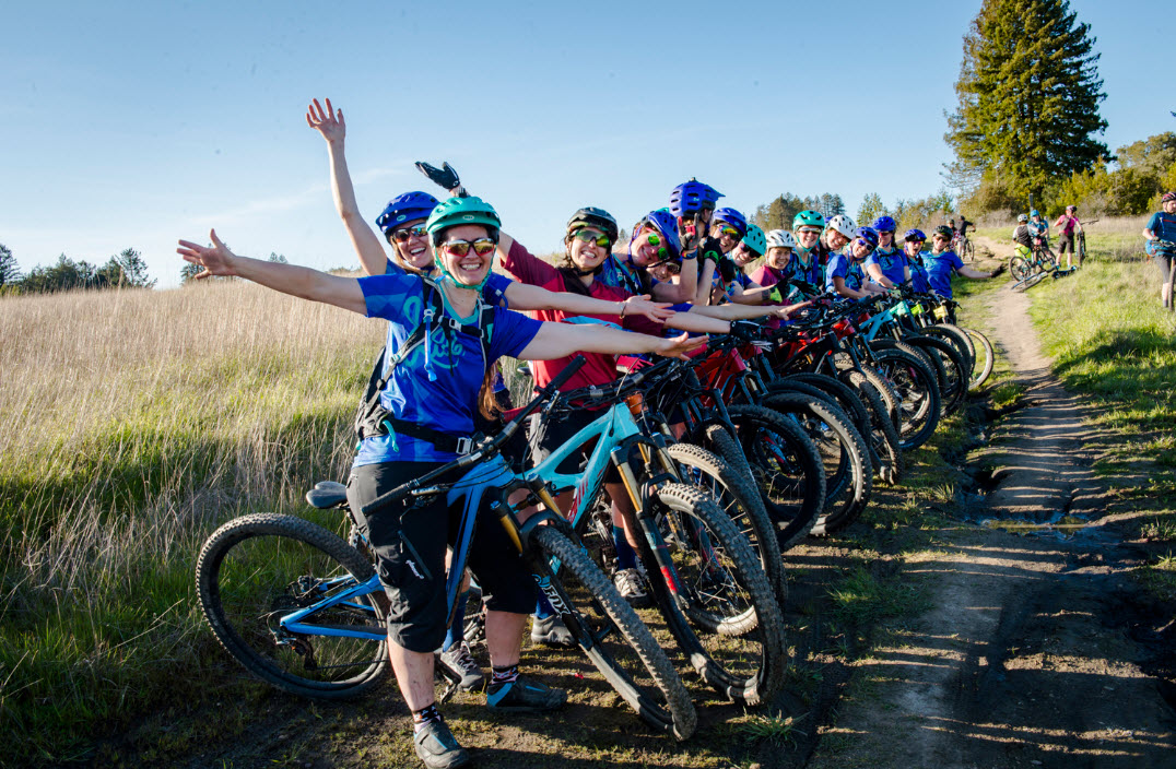 Many people on bicycles lined up on the side of a dirt road.