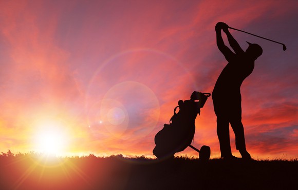 A golfer swings next to his bag at sunset. 