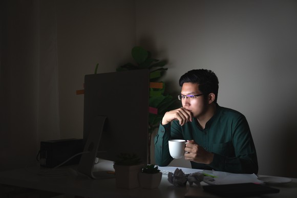 An Asian man sitting at his laptop in the dark, holding a mug of tea or coffee