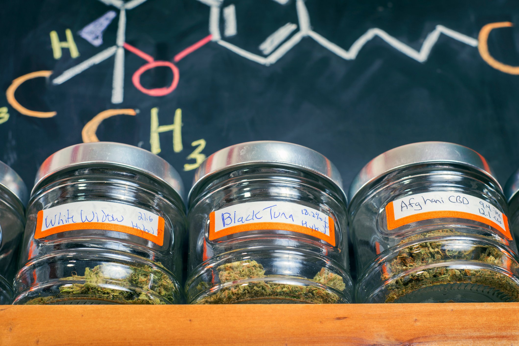 Three jars of dried cannabis flowers sitting on a ledge against a blackboard.