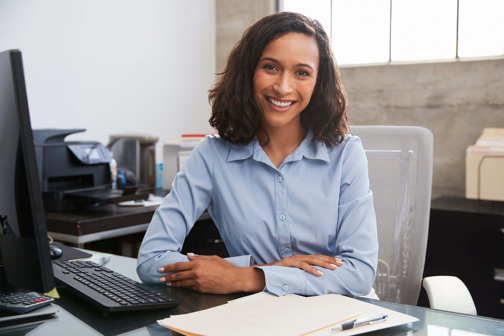Woman in dress shirt sitting at desk, smiling with arms crossed