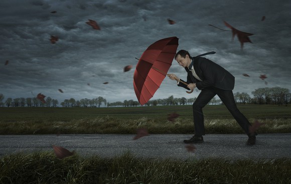Man in suit pushing against wind with umbrella, with dark clouds in the background.