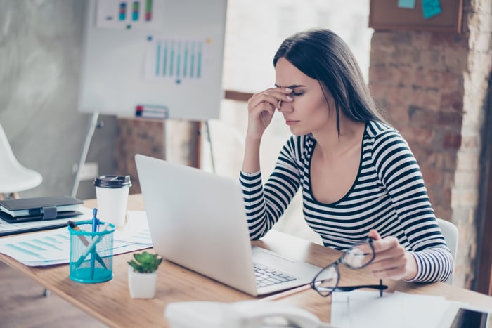 Woman sitting at desk in front of laptop, holding eyeglasses in one hand and pressing fingers against her nose with the other