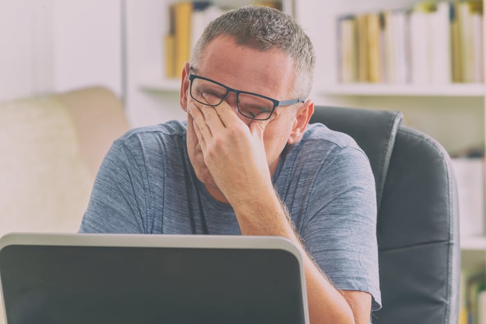 Bespectacled man at his laptop, covering his eyes with his left hand. 