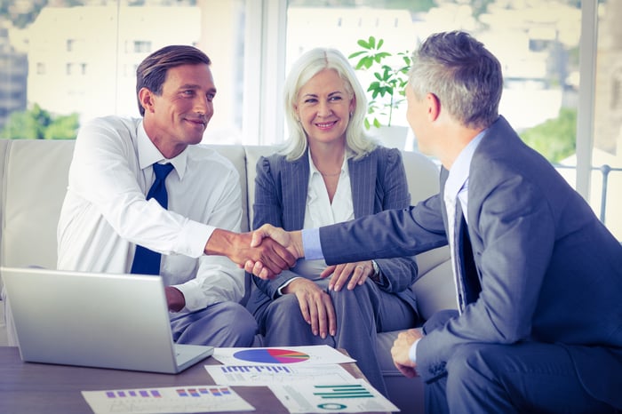 Two men and a woman in business attire sitting in front of a laptop and some paperwork; the men are shaking hands