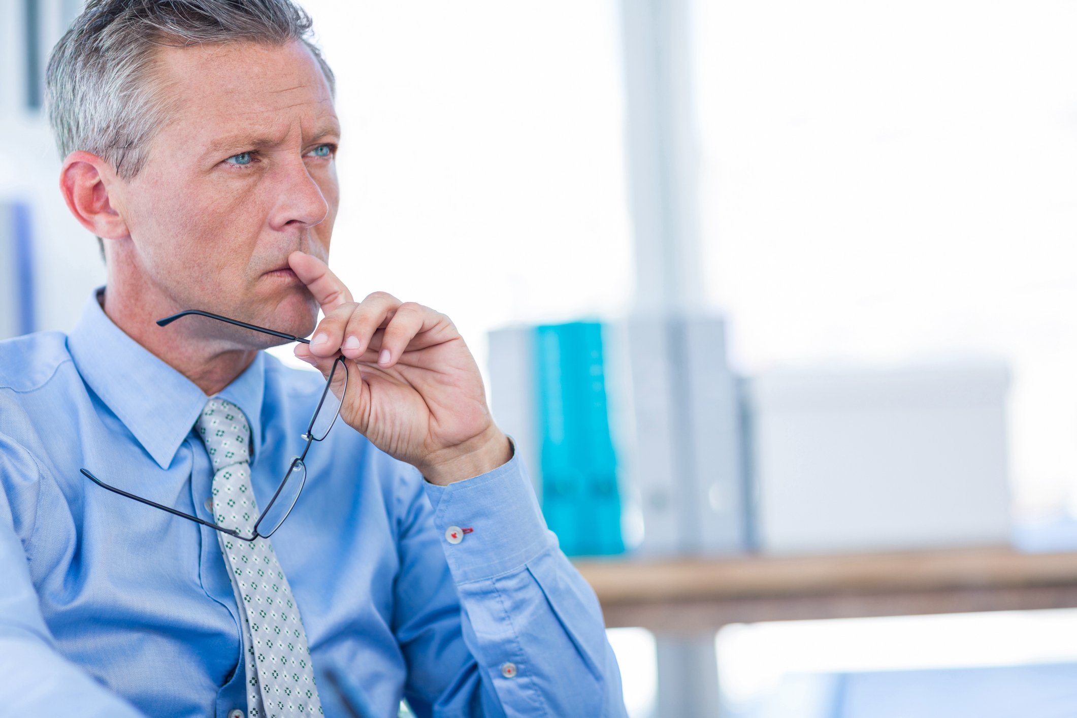 Older man in dress shirt and tie holding a pair of eyeglasses and putting his finger to his mouth as if deep in thought