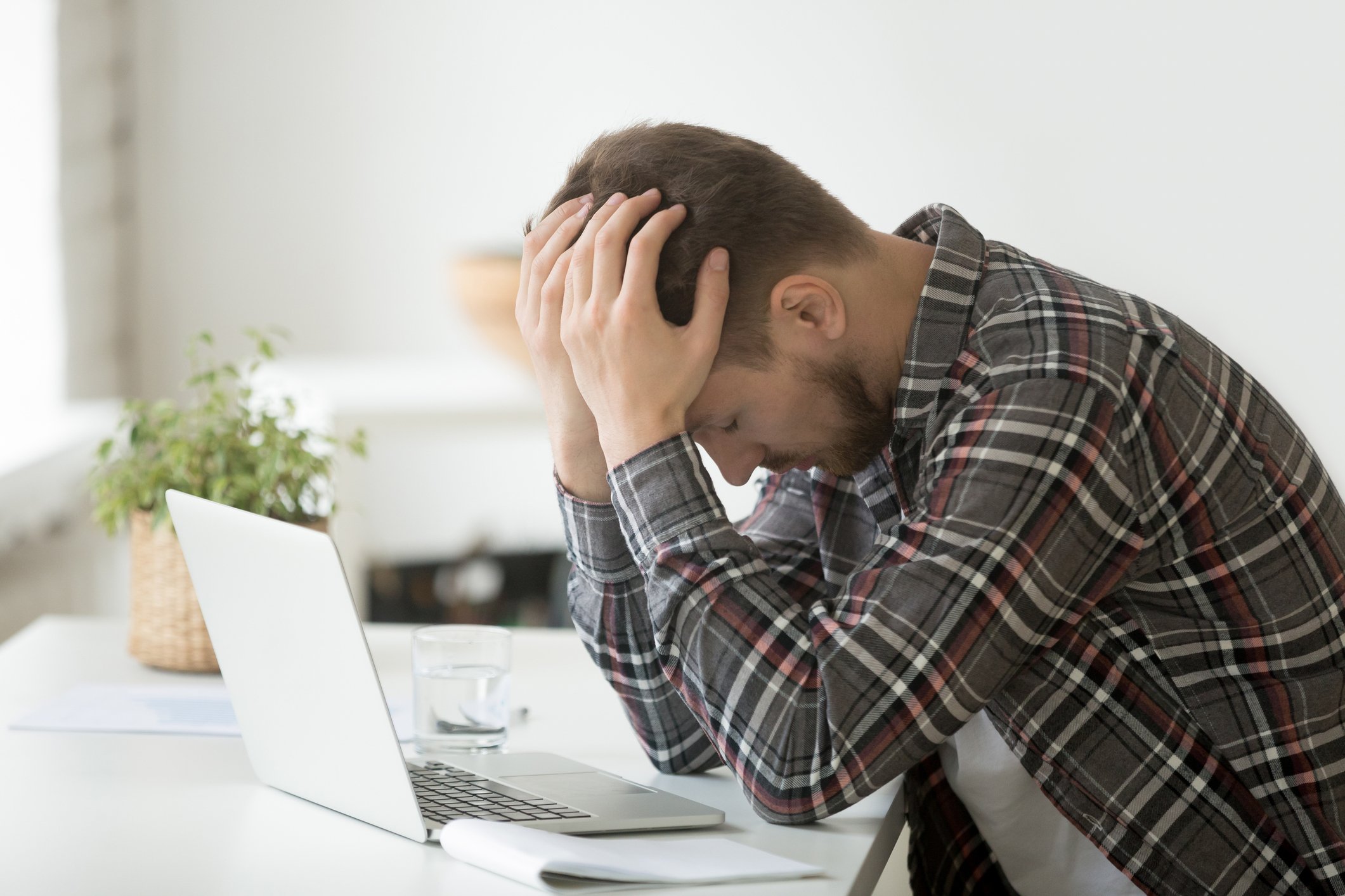 A man in a plaid shirt puts his head in his hands in anguish sitting in front of his laptop. 