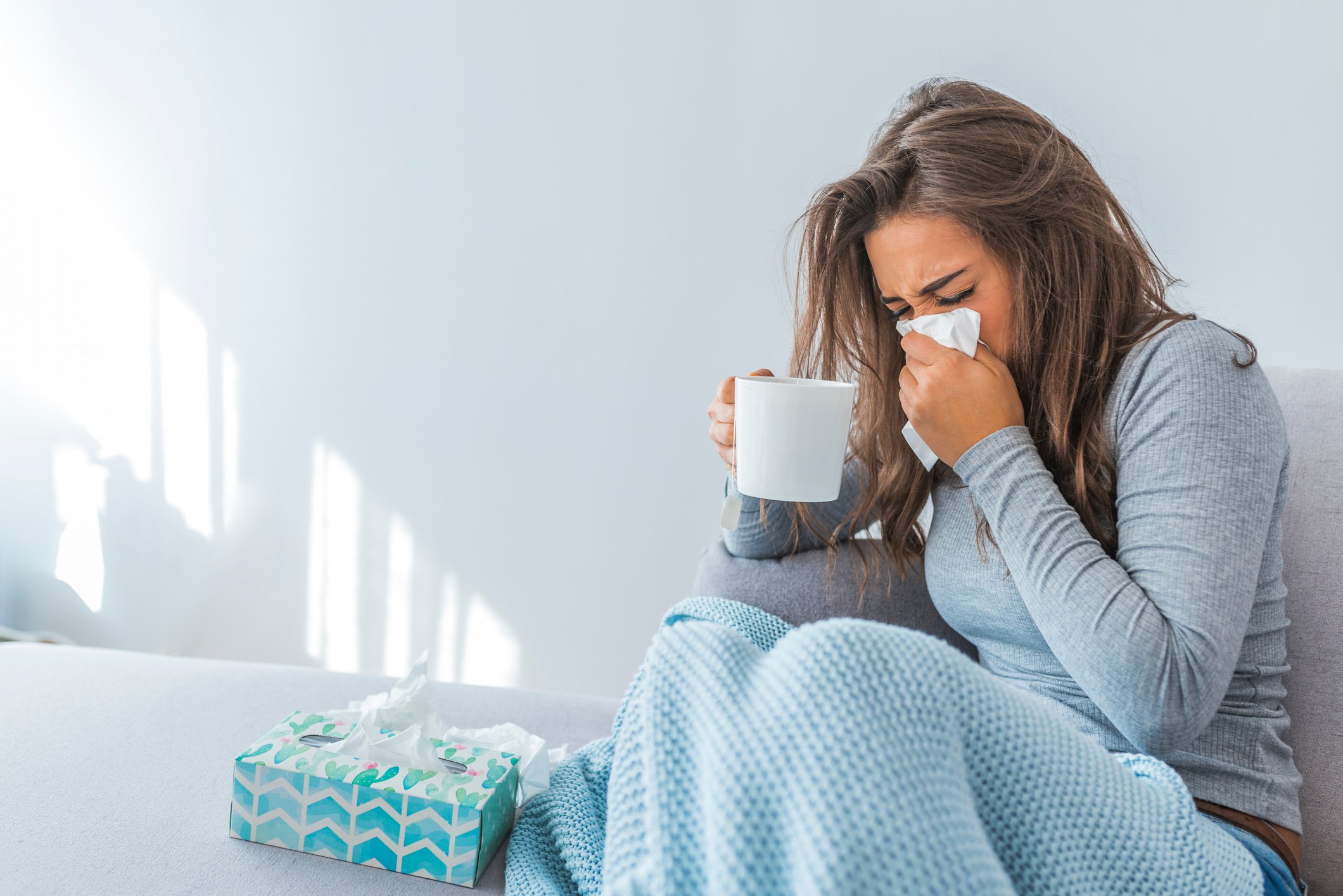 Woman sitting under a blanket on a bed and holding mug in one hand and blowing nose with other hand, with a box of tissues by her side.