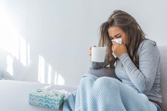 Woman sitting under a blanket on a bed and holding mug in one hand and blowing nose with other hand, with a box of tissues by her side.
