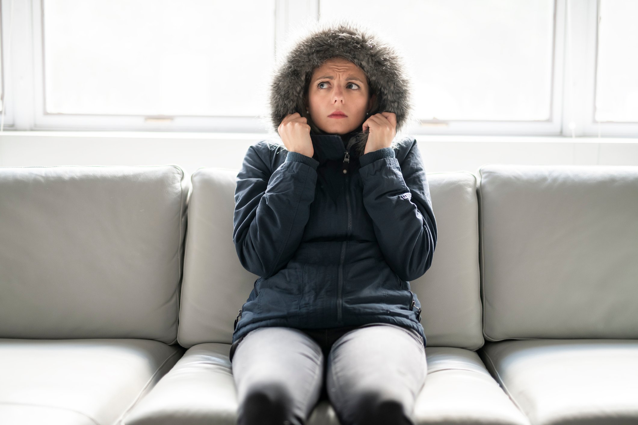 A women in a heavy coat and a worried look sits on a couch.