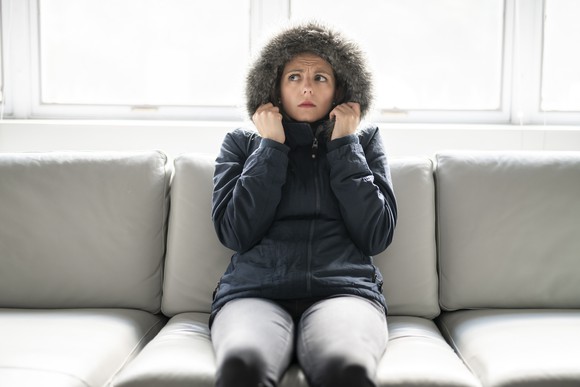 A women in a heavy coat and a worried look sits on a couch.