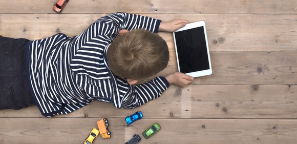 A boy laying on the floor using a tablet computer with little toy cars laying around him.