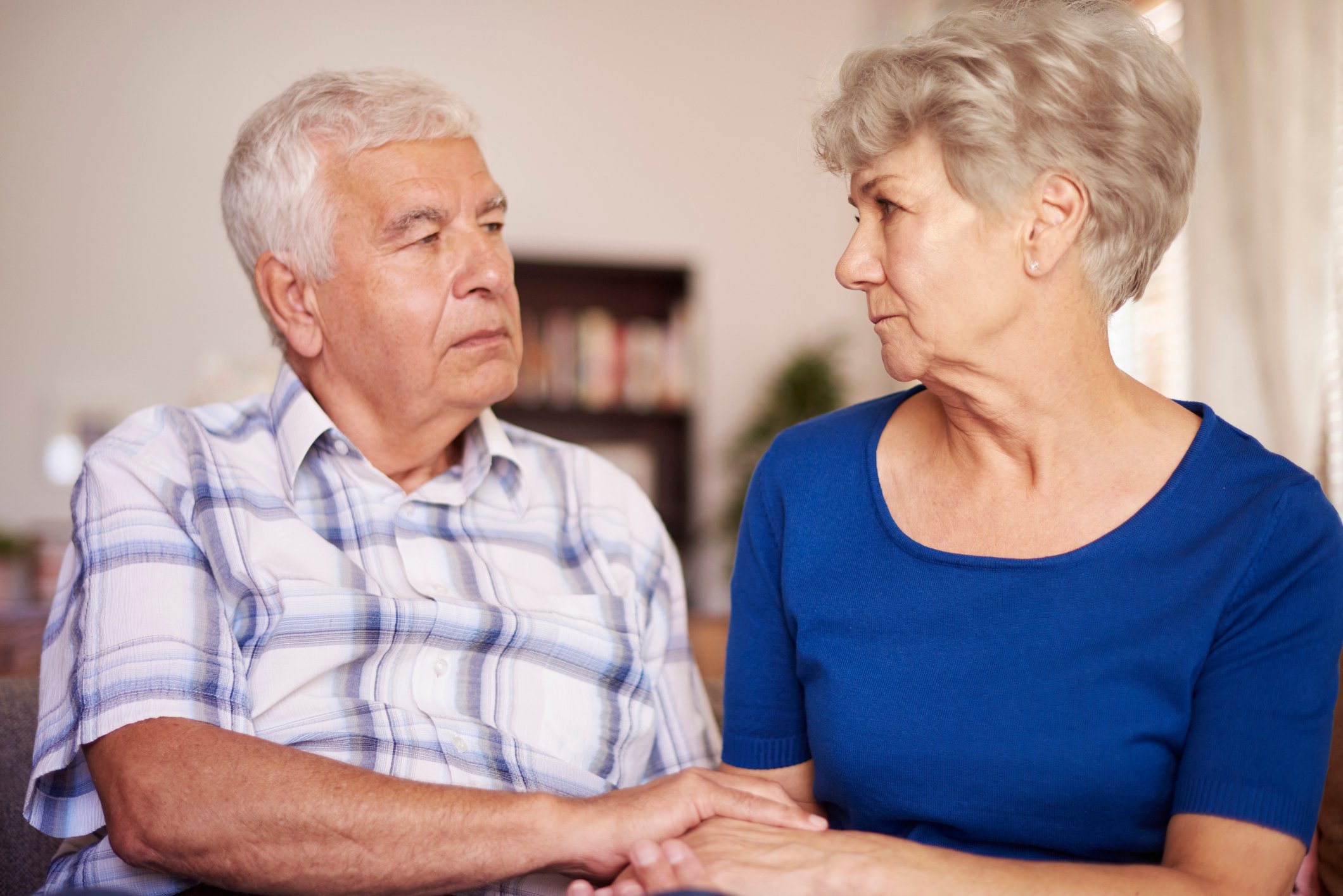 Senior man and senior woman looking at each other with serious expressions and holding hands.
