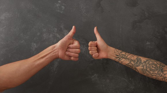 One with tattoos and one arm without tattoos give the thumb's up sign.